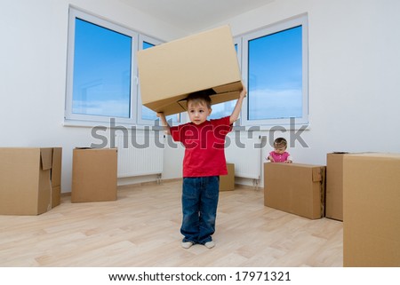 A little boy moving cardboard boxes in his new house, while his little sisters plays in the background.