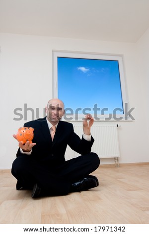 A business man sitting on the floor holding up a piggy bank after he has saved up for a new home.