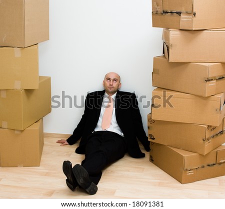 Man sitting on a floor against a wall, with tall stacks of cardboard boxes on each side of him.