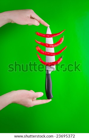 A view of fingers holding a kitchen knife with several whole red peppers on it.  Green background.