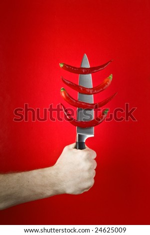 Hand holding a carving knife with a row of red chili peppers on the blade against a red background.