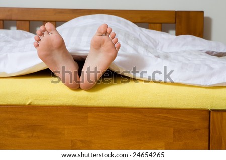A closeup view of two bare feet sticking out from under a blanket at the end of a bed.