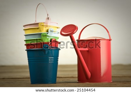 Six colored buckets in column with red watering can on timber floor