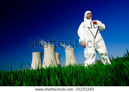 A view of a man in a protective hazardous material suit, standing in front of a nuclear power plant, holding a white arrow.