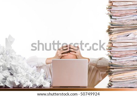 stock photo : A stressed person holding his head behind a laptop surrounded by a pile of files and papers,