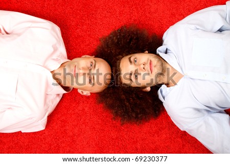 Two young men laying on a red carpet, one is bald and the other has a full head of hair.