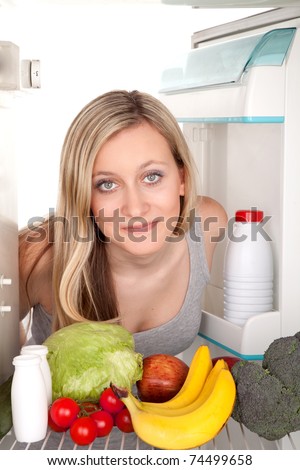 Smiling teenage girl looking inside of a refrigerator with fresh fruits and vegetables.