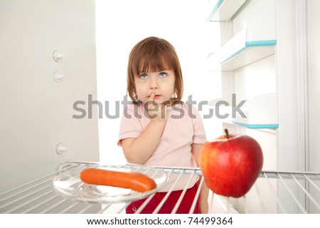 Cute young girl looking in open refrigerator deciding between healthy apple and unhealthy hot dog.