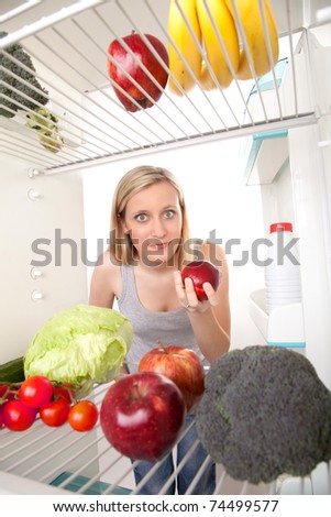 Young woman holding an apple and looking into a refrigerator filled with fruits and vegetables.