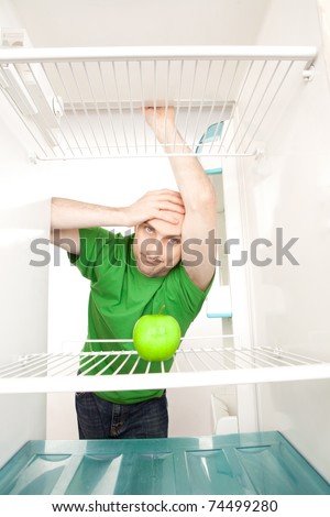 Young man leaning in open doorway of open refrigerator with single apple on empty shelves.