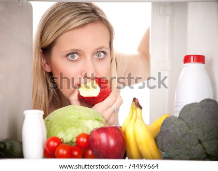 Attractive female teenager eating apple and looking at healthy fruit and vegetables in refrigerator.
