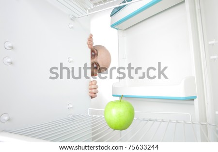 Bald head of young man looking around corner of open refrigerator containing single green apple.