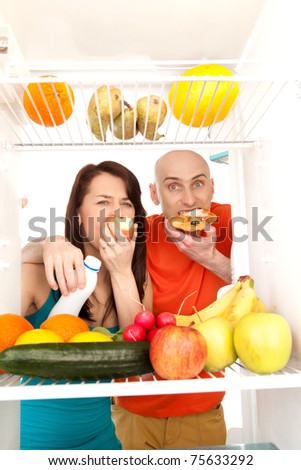 A couple eating cream cakes standing in the open door of the refrigerator full of healthy fresh fruit.