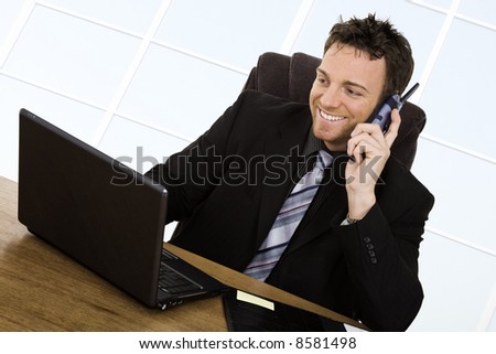 Caucasian businessman setting at a desk talking on the phone and smiling while working on a laptop