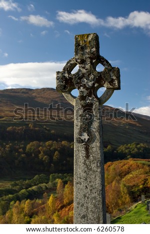 A Celtic Cross in a Scottish Highland graveyard, with autumn coloured trees in the background.