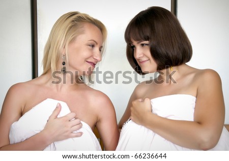 Two attractive naked women covered with white sheet, sitting in bed and looking at each other
