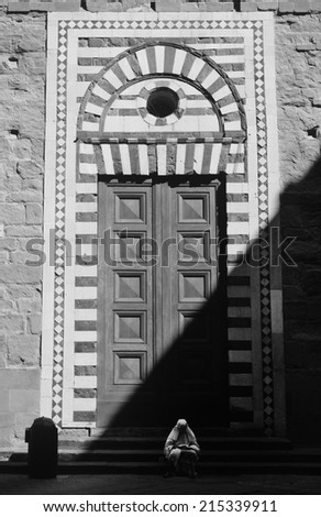 Figure in white sitting and holding bread, in front of church doors, which are in half shadow, no recognizable person, Black and White photo, small amount of noise may occur