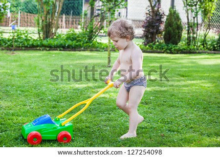 Little blond toddler boy playing with lawn mower machine in summer garden on sunny day