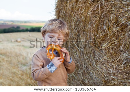 Cute toddler eating German pretzel on golden hay field in summer.
