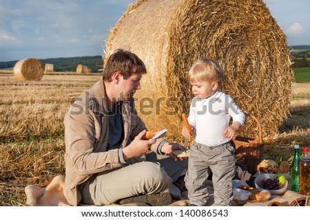 Young man and little toddler boy making evening picnic on hay field, Germany.