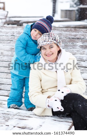Portrait of young mother and her adorable son sitting in winter on bench with snow