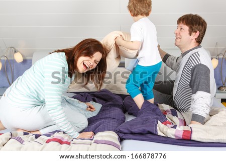 Happy family of a three having fun in pajamas at home in bed and fighting with pillows