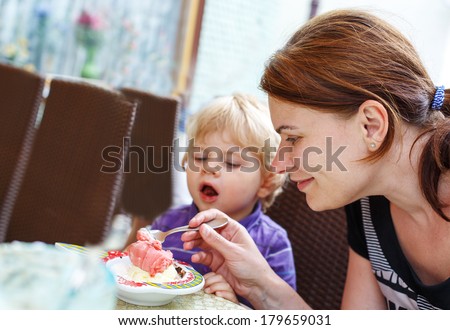 Young mother feeding her little boy son with ice-cream in city cafe in summer.