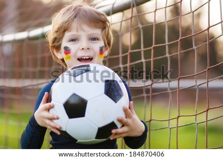 Funny little boy of 4 having fun with playing soccer with football on football field, outdoors. In goalkeeper uniform and painted German flag on face.