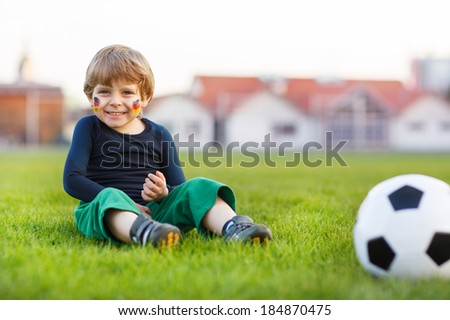 Funny little boy of 4 having fun with playing soccer with football on football field, outdoors. In goalkeeper uniform and painted German flag on face.