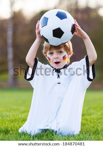 Funny little boy of 4 having fun with playing soccer with football on football field, outdoors. In white German national uniform and painted German flag on face.