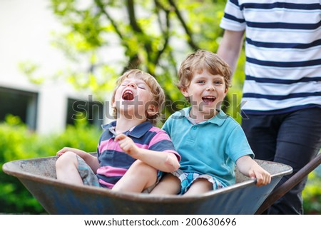 Two little siblings having fun in a wheelbarrow pushing by father in summer garden