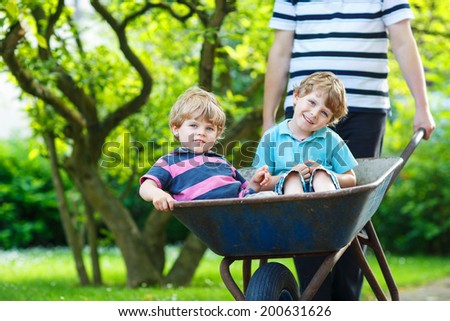 Two little boys having fun in a wheelbarrow pushing by father in summer garden