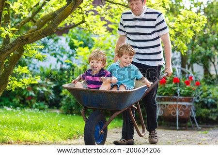 Two little children having fun in a wheelbarrow pushing by father in summer garden