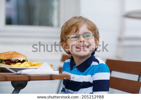 Cute little boy eating fast food: french fries and hamburger in cafe