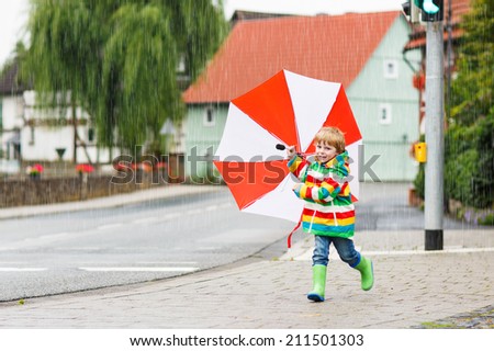 Little child having fun with red umbrella, wearing colorful jacket outdoors at rainy day