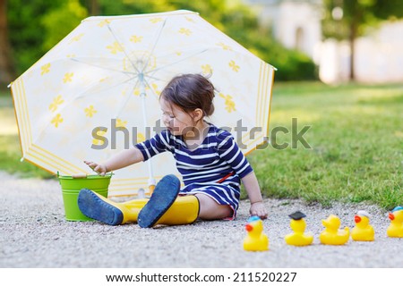 Cute toddler girl having fun with umbrella in yellow rain boots and rubber ducks in summer park.