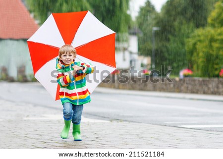 Funny boy having fun with red umbrella, wearing colorful raincoat and green boots outdoors at rainy day