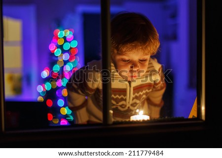 Little cute boy standing by window at Christmas time and blowing candle. With colorful lights from Christmas tree on background, selective focus.