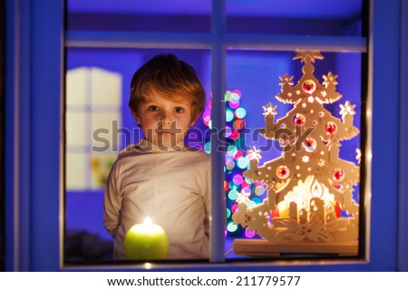 Little boy standing by window at Christmas time and holding candle. With colorful lights from Christmas tree on background, selective focus.