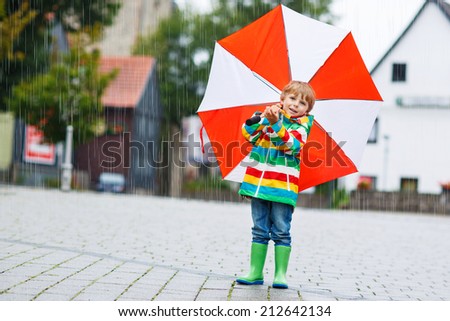 Smiling little boy walking in city with red umbrella, wearing colorful rain coat and green boots outdoors at rainy day