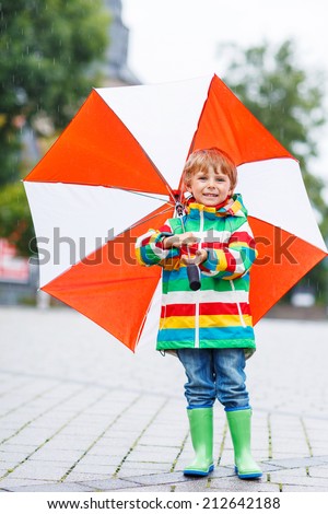 Cute smiling little boy walking in city with red umbrella, wearing colorful rain coat and green boots outdoors at rainy day