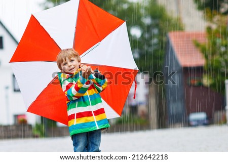 Smiling little boy walking in city with red umbrella, wearing colorful rain coat and green boots outdoors at rainy day