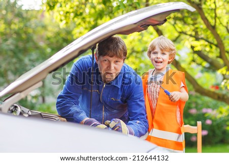 Young dad in overal teaching his little boy in safety vest  to repair motor oil in family car.