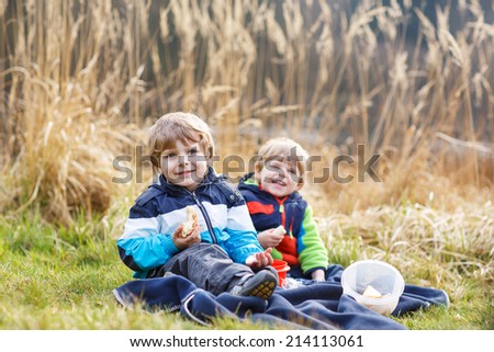 Two little sibling boys having picnic near forest lake, nature on cold spring or autumn day.