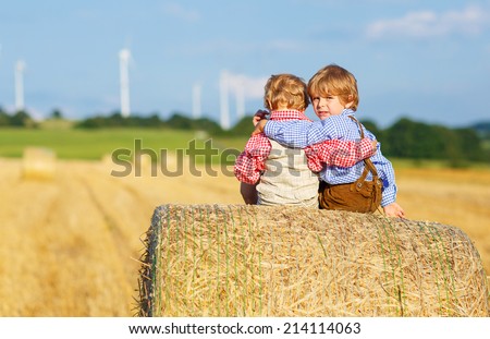 Two little sibling boys and friends in traditional German bavarian clothes sitting on hay stack or bale and speaking on yellow wheat field in summer
