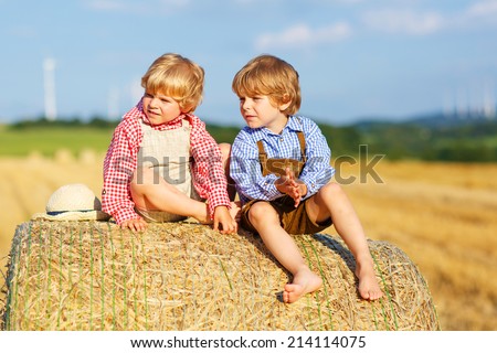 Two little friends in traditional German bavarian clothes sitting on hay stack or bale and speaking on yellow wheat field in summer