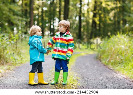 Little twin boys in colorful waterproof raincoats and rubber boots walking through autumn forest together.