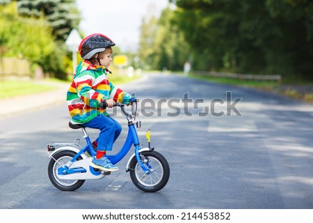Adorable kid boy in red safety helmet and colorful raincoat riding his first bike on summer day.