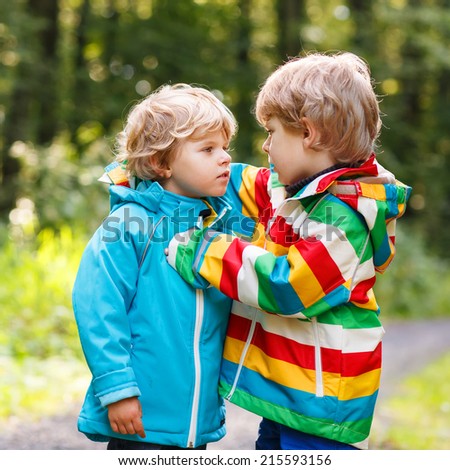 Two little sibling boys in colorful waterproof raincoats and rubber boots hugging in autumn forest together. Brother love