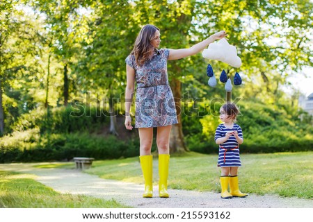 Young mum and her little toddler girl in yellow rubber boots, family look, in summer park playing with rain drops toy